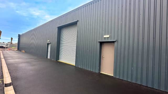 Gray metal building with a large roller door and two smaller doors, set on a freshly paved driveway.
