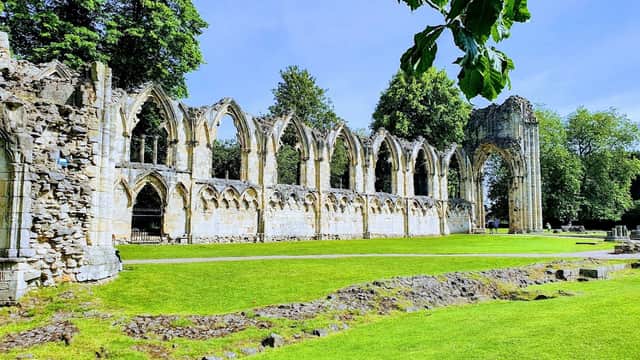 Historic stone ruins with arched windows, surrounded by lush green grass and tall trees under a clear blue sky.