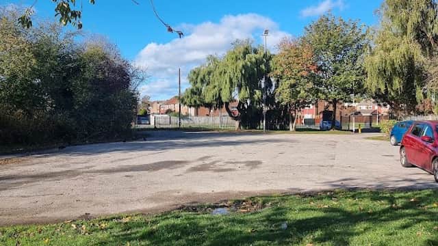 Alderman Kneeshaw Car Park with a clear blue sky, green grass, and trees surrounding a gravel parking area.