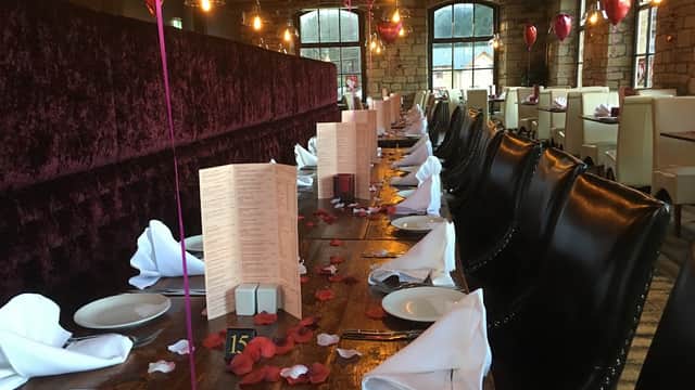 Elegant dining setup with dark velvet seating, white table settings, and rose petals on a wooden table in a cozy atmosphere.