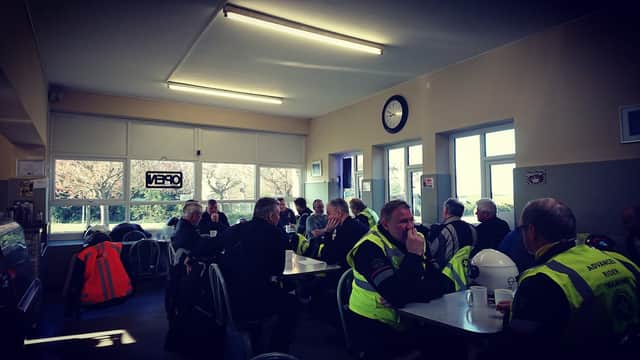 Busy cafe interior with patrons in high-visibility jackets enjoying food and drinks at tables, bright windows letting in light.