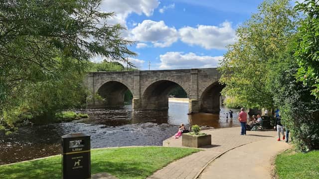 Wilderness Car Park by a river, with a stone bridge, trees, and people enjoying the scenic view on a sunny day.