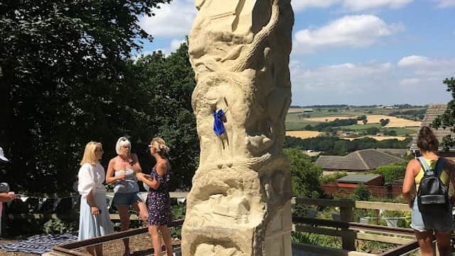 A tall stone sculpture surrounded by visitors, with rolling hills and a blue sky in the background.