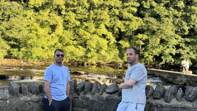 Two men stand by a stone wall near a river with lush green trees in the background, enjoying a sunny day.