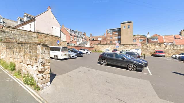 Car park with multiple vehicles, stone walls, and buildings under a clear blue sky in Whitby, Yorkshire.