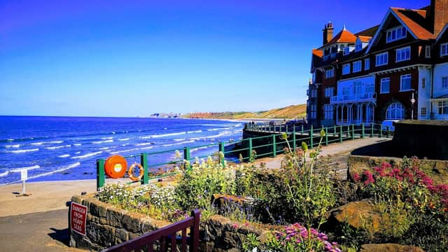 Sandsend beachside view with a car park, vibrant flowers, and a seaside building under a clear blue sky.