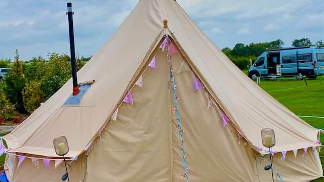 A beige bell tent adorned with bunting, set on green grass with a van parked nearby under a cloudy sky.