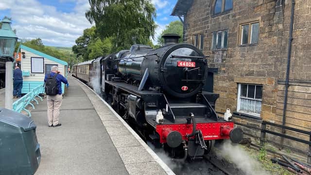 Steam train at Grosmont Station, with passengers waiting on the platform and historic stone buildings in the background.