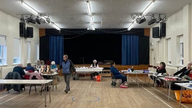 Spacious village hall interior with tables, chairs, and a stage in the background, set up for an event.