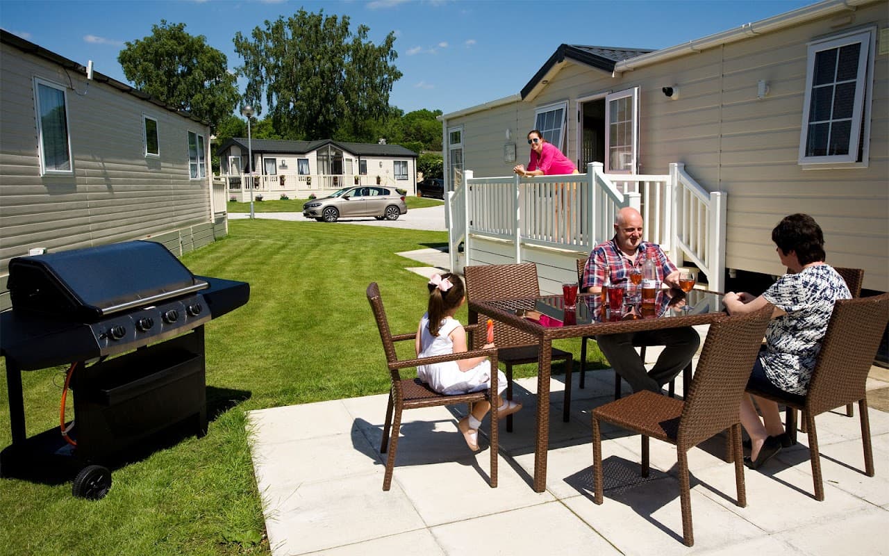 A family enjoying drinks at a patio table near a grill, with holiday homes and green lawns in the background.