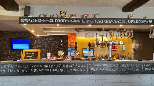 Bar counter with taps, beer glasses, and a menu board; cozy interior with wooden beams and decorative wallpaper.