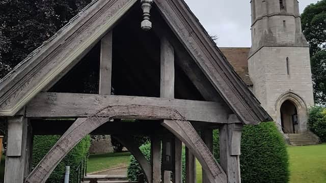 Wooden entrance structure leading to a stone church tower amidst lush greenery and a cloudy sky.