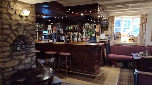 Cozy bar interior with wooden counter, stone wall, stools, and warm lighting, featuring shelves of drinks and a plaid carpet.