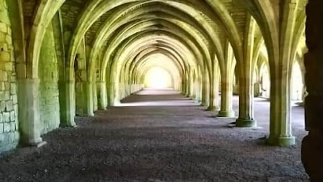 Arched stone columns create a shadowy corridor leading to bright light at the end, surrounded by textured walls.