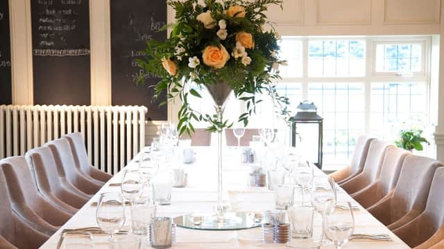 Elegant dining setup with a long table, glassware, and a tall floral centerpiece featuring roses and greenery.
