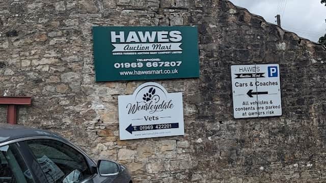 Hawes Farmers' Auction Mart sign on a stone building, with parking and directional signs for cars and coaches.