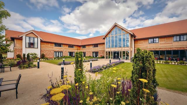 Modern restaurant with large windows, surrounded by landscaped gardens and colorful flowers under a partly cloudy sky.