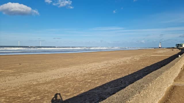 A sandy beach with gentle waves, distant wind turbines, and a clear blue sky with scattered clouds.