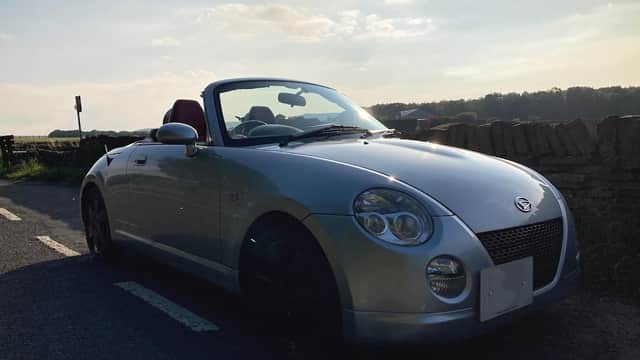 Silver convertible car parked on a country road with a scenic backdrop of hills and a blue sky.