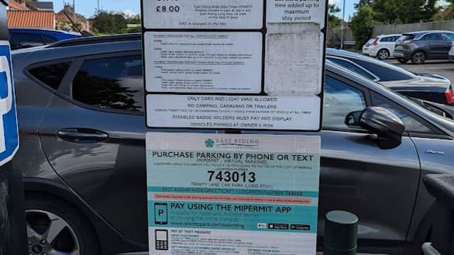 Signboard displaying parking tariffs and payment options at Trinity Lane Car Park, with cars parked nearby.