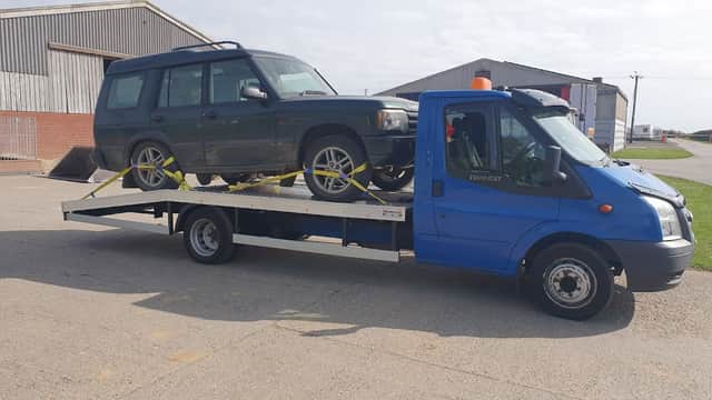A blue tow truck carrying a green vehicle on a flatbed, with storage buildings in the background.