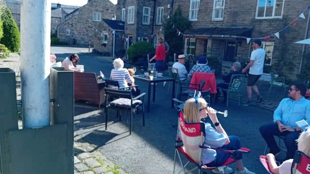 People socializing outside at Burton Leonard Cricket Club, with chairs, tables, and festive bunting decorating the area.