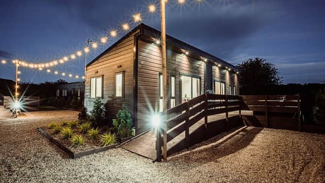 Modern wooden building with outdoor lighting, surrounded by gravel and greenery, set against a twilight sky.