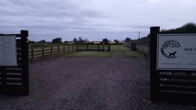 Entrance to South Acres Dog Fields with wooden gates, gravel path, and open grassy fields under a cloudy sky.