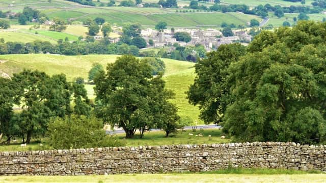 View of lush green hills, trees, and a stone wall in Askrigg, Yorkshire, with a distant village in the background.
