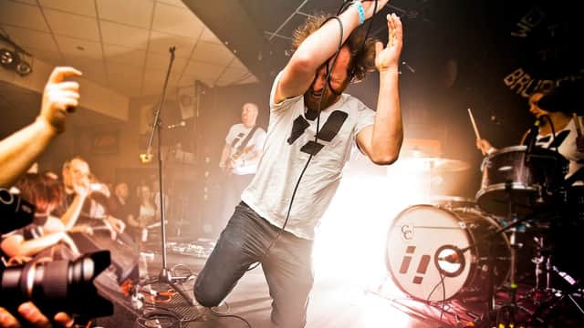 A performer kneels on stage, singing passionately, with a lively crowd and band in the background.