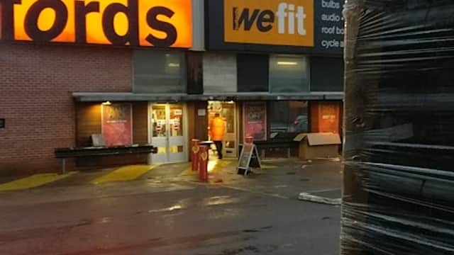 Halfords store entrance with bright signage, wet pavement, and stacked pallets in foreground.