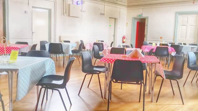 Brightly lit hall with tables set in blue and red checkered cloths, surrounded by black chairs and wooden flooring.