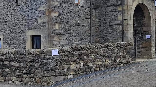 Stone barn with a large archway entrance, surrounded by a low stone wall and gravel path, set against a cloudy sky.
