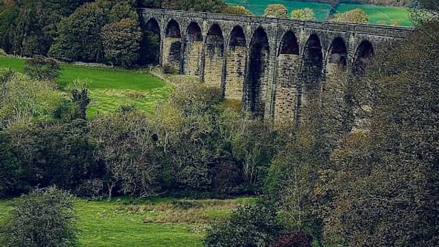 Hewenden Viaduct spans lush green fields, framed by trees under a dramatic cloudy sky in Cullingworth, Yorkshire.