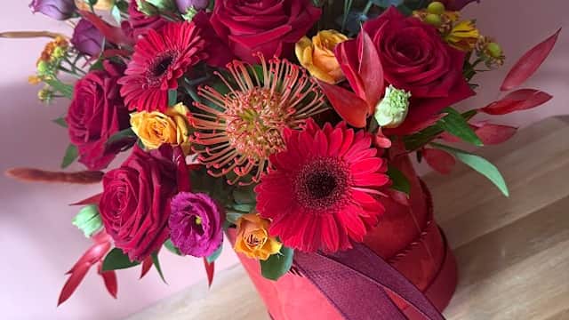 Vibrant floral arrangement in a red bucket, featuring roses, gerberas, and tropical blooms against a pink background.