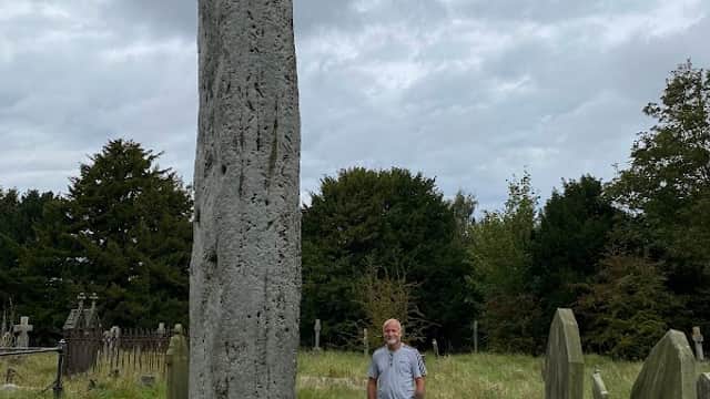 Tall, weathered stone monolith surrounded by grass and gravestones, with a person standing nearby under a cloudy sky.