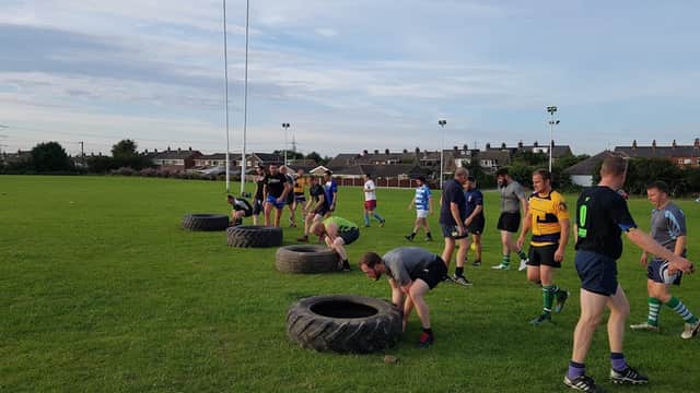 Rugby players training on a grassy field, using large tires for drills, with goalposts in the background.