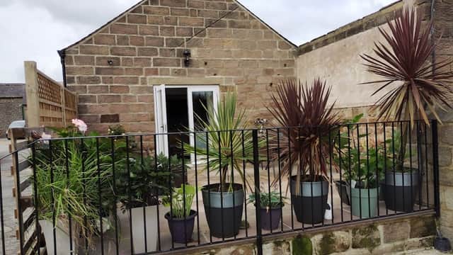 A stone building with a patio surrounded by potted plants and a black railing in a serene outdoor setting.