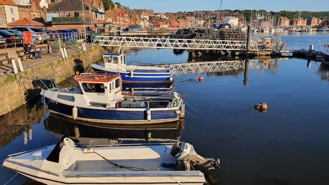 Boats moored at a dock in Whitby, Yorkshire, with buildings and hills in the background under a clear blue sky.