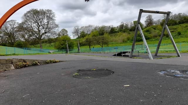 Empty car park with a swing set and a large orange structure, surrounded by green hills and cloudy skies.
