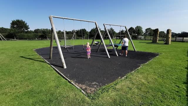 A sunny playground with swings, a child playing, and a grassy area surrounded by trees in Borrowby, Yorkshire.