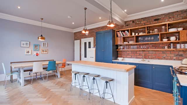 Modern kitchen with blue cabinetry, wooden dining table, colorful chairs, and exposed brick wall. Stylish lighting fixtures.