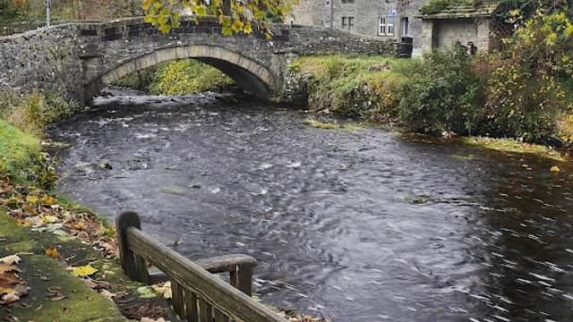 A stone bridge over a flowing river, surrounded by autumn leaves and historic buildings in Clapham, Yorkshire.