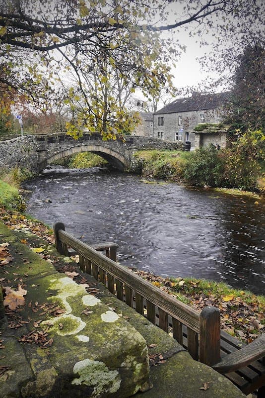 A stone bridge over a flowing river, surrounded by autumn leaves and historic buildings in Clapham, Yorkshire.