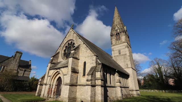 Victorian church with a tall spire, large rose window, and stone facade, surrounded by green grass and trees.