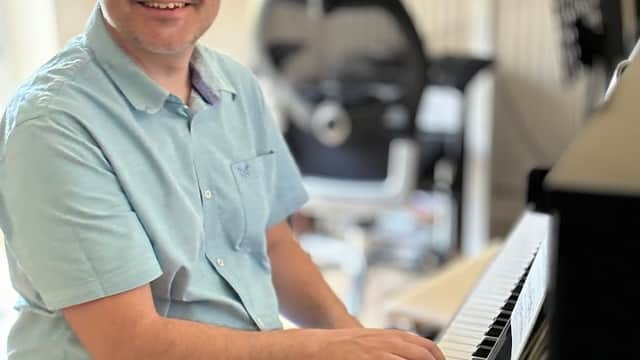 A man with glasses smiles while playing a piano in a music shop setting with chairs and music stands in the background.