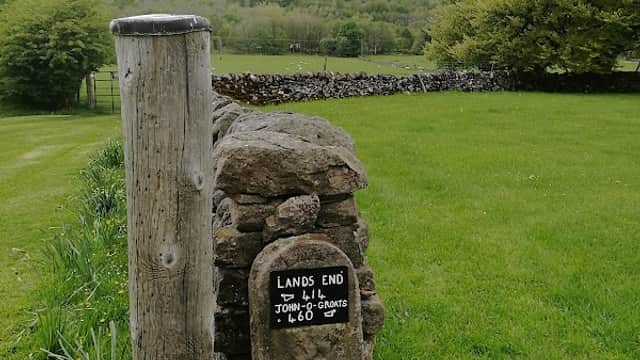 Signpost reading "Lands End" next to a stone pillar, with lush green grass and hills in the background.