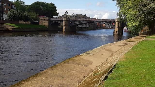 View of a riverbank in York with a bridge, greenery, and a cloudy blue sky above.