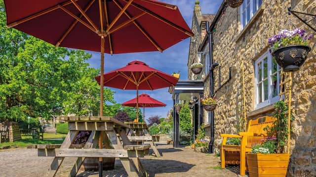 Stone building with red umbrellas over wooden tables, flowers in hanging baskets, and a sunny outdoor seating area.