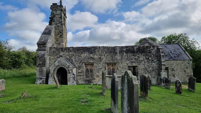 Ruined church with a tower, surrounded by gravestones and lush greenery under a cloudy sky.